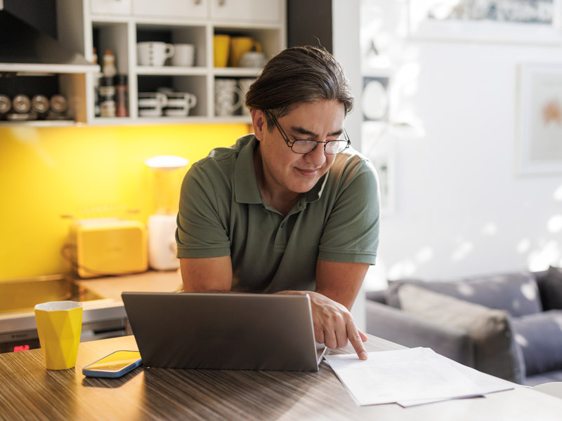 Image of man reading sheet of paper while on laptop